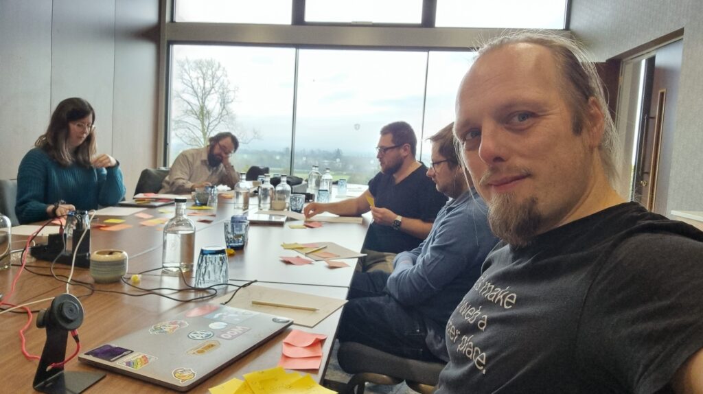 Dan sits at a boardroom table in an airy, bright room. He's wearing an Automattic t-shirt that reads "Let's make the Web a better place." In the background, several other people discuss a pile of post-it notes that have begun to pile up on the table.