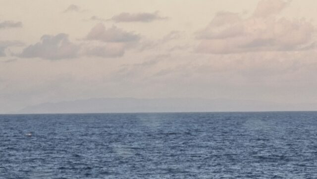 Across the sea, a rugged island begins to emerge over the horizon, through distant clouds.