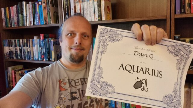 Dan, in a library, smiles as he holds up a certificate that certifies that he is an Aquarius.
