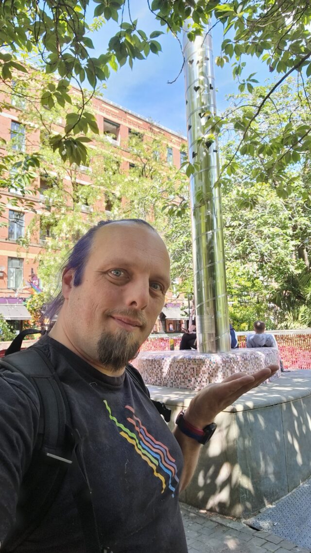 Dan, wearing a pride rainbow t-shirt, stands alongside a steel beacon in the corner of an urban park, under bright late-spring sunlight.