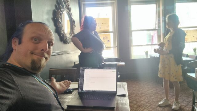 Dan gestures at his laptop in a quiet pub function room, on which he's writing some code. In the background, two women are having a conversation.